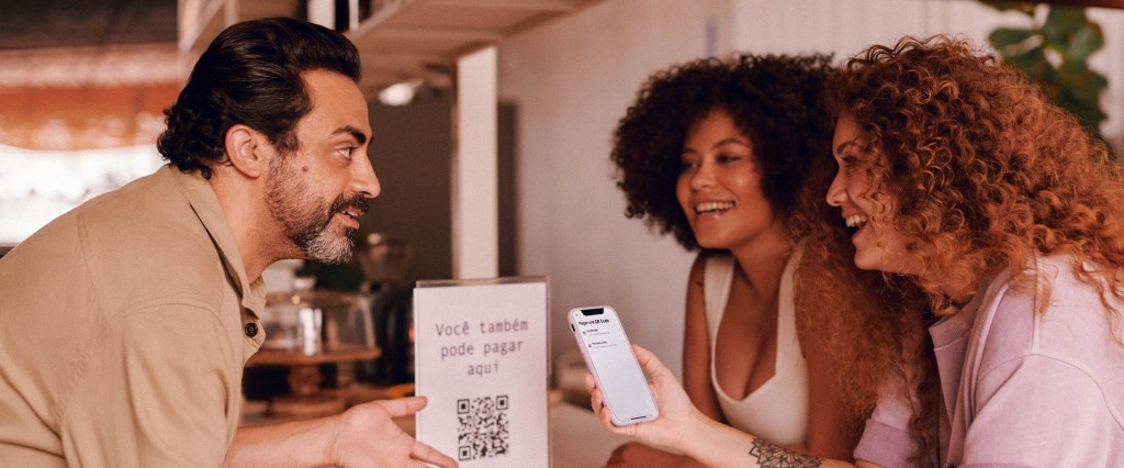 Na imagem aparece um homem de meia idade do lado esquerdo, atrás do balcão de seu estabelecimento comercial, vestindo uma roupa em tons de bege com os cabelos curtos e barba. Ele olha para duas mulheres que estão do lado direito do balcão, uma é negra e usa uma camiseta branca de alça e a outra é ruiva usa uma camiseta em tom roxo claro e segura um celular