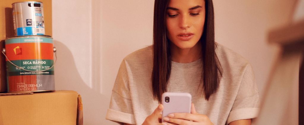 Foto de uma mulher sentada com um celular na mão. Ela tem cabelo liso e comprido e está usando uma camiseta cinza.