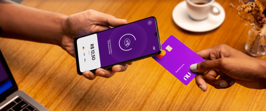 Nubank lança NuTap: na foto, vemos duas mãos - uma segurando um celular, onde é possível visualizar a tela de cobrança do NuTap, e outra segurando um cartão do Nubank, simulando um pagamento por aproximação.