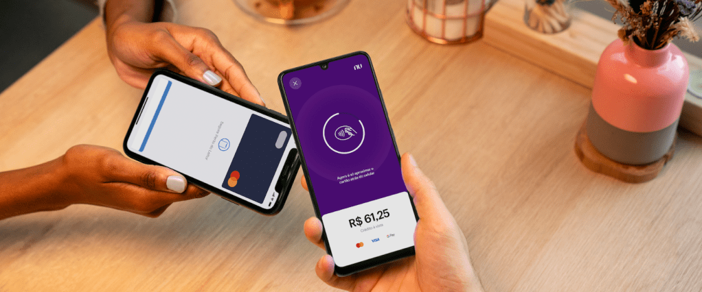 NuTap: na imagem, é possível visualizar um pagamento sendo feito com o NuTap. Uma mão segura um celular onde aparece a tela de cobrança do app. Outra, segura outro celular com carteira virtual, fazendo um pagamento por aproximação.