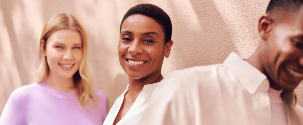 Foto de três pessoas em frente a um muro branco, todas sorrindo: uma mulher no fundo usa uma blusa lilás, uma mulher no meio e em mais destaque está com uma roupa clara com jeito mais executivo e um homem de camisa clara está quase saindo da cena.