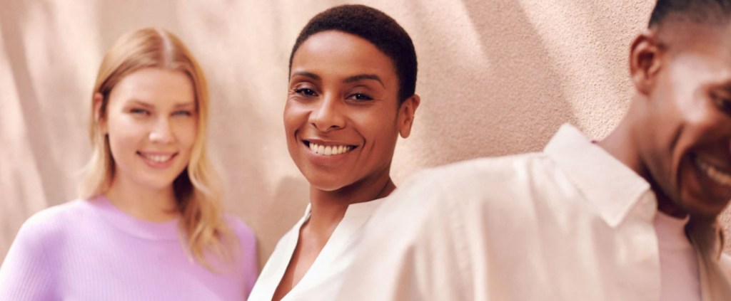 Foto de três pessoas em frente a um muro branco, todas sorrindo: uma mulher no fundo usa uma blusa lilás, uma mulher no meio e em mais destaque está com uma roupa clara com jeito mais executivo e um homem de camisa clara está quase saindo da cena.