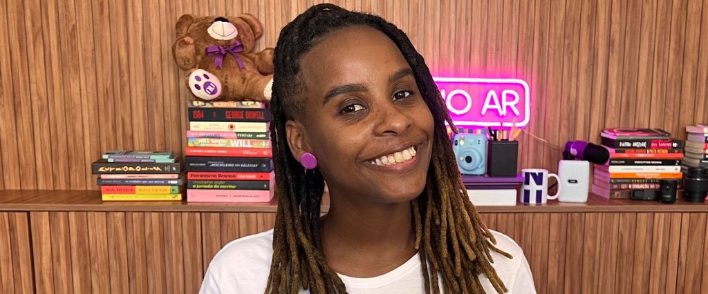 Monique Evelle sorridente para a foto, veste uma camiseta branca. Ao fundo existe uma estante de madeira com diversos acessórios, como livros, um letreiro luminoso roxo escrito no ar e um ursinho.