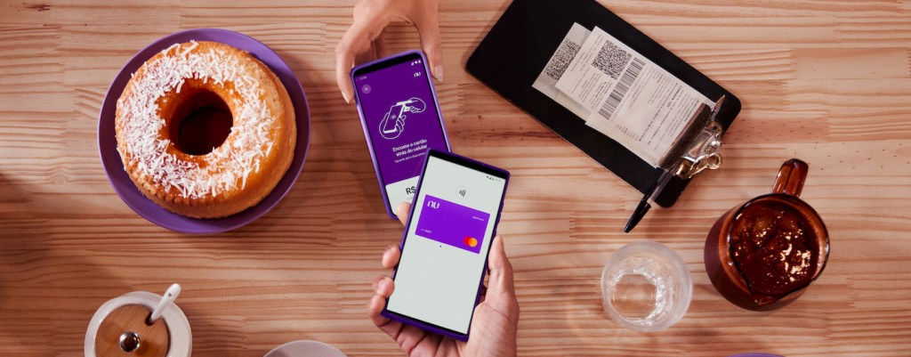 Tap to Pay: tudo sobre a maquininha de cartão no app do Nubank. Imagem mostra uma mesa de madeira clara com: um bolo redondo de coco em um prato roxo, um açucareiro branco com tampa de madeira, uma xícara de café cinza e seu respectivo pratinho. Ao lado direito da imagem , a mão de uma pessoa branca segurando um celular na tela de cobrança do 