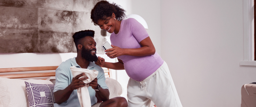 Buscador de boletos do Nubank: na imagem, um homem e uma mulher negros estão sorrindo enquanto olham olham para um celular branco na mão dela. A mulher está grávida, com a barriga aparente, e em pé ao lado do homem, que está sentado na cama.