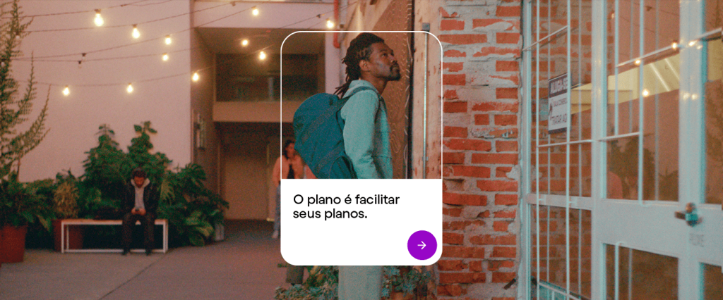 Foto mostra um homem negro, com barba e dread, vestido de verde, com uma mochila, esperando o elevador num lugar que tem paredes de tijolinhos.