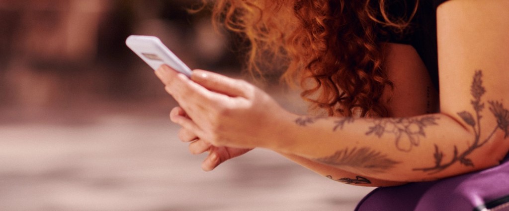 Na foto, os braços tatuados de uma mulher ruiva que segura um celular.