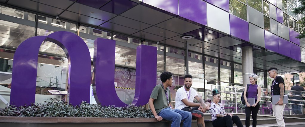 Imagem da fachada do escritório do Nubank, em São Paulo. Em frente ao prédio está o logo do NU bem grande, e funcionários estão sentados junto a ele.