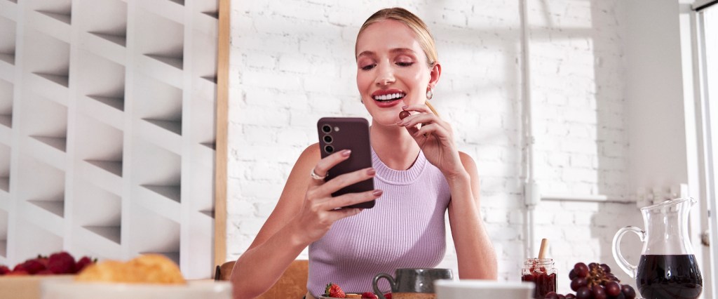 Imagem de uma mulher em uma sala de jantar, tomando café da manhã enquanto segura o celular. Ela é branca, loira, e veste uma camiseta lilás. A mesa tem frutas, suco e xícaras de porcelana.