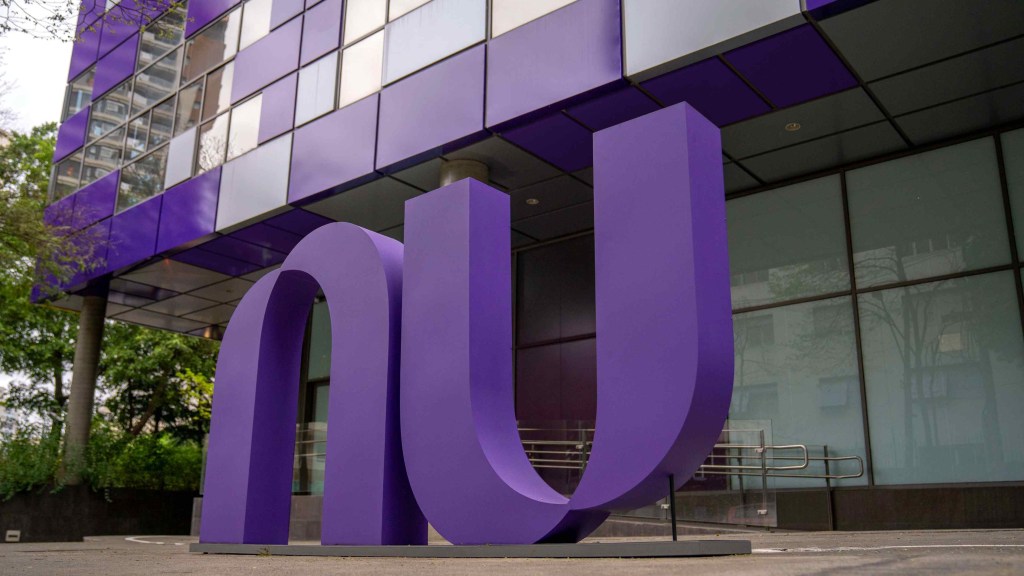 Na imagem aparece o logo do Nubank em roxo em frente ao escritório, que combina vidro e partes roxas