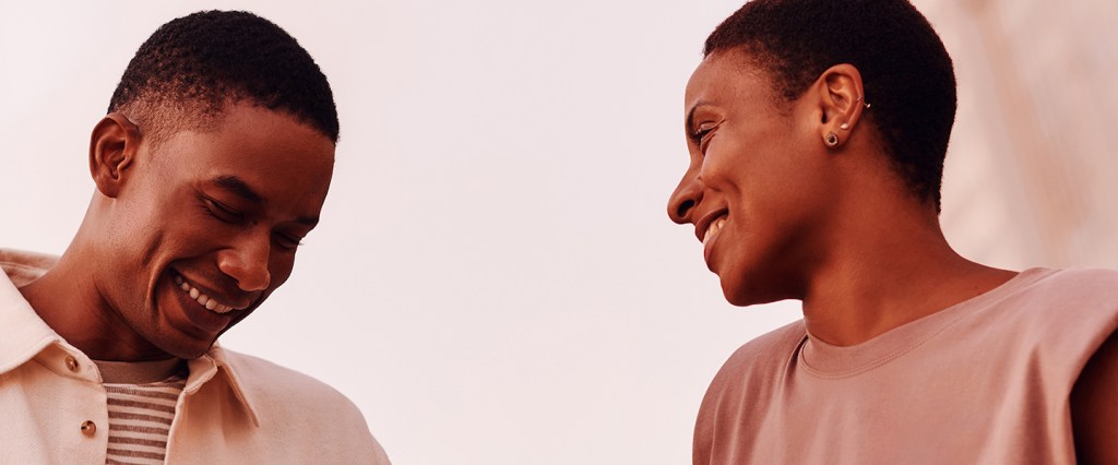 Um homem negro e uma mulher negra estão sorrindo. Ele olha para baixo e ela olha para ele. Ambos tem cabelo raspado e vestem roupas claras, em tons de bege.