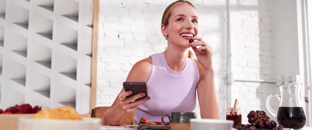 Imagem de uma mulher em uma sala de jantar, tomando café da manhã enquanto segura o celular. Ela é branca, loira, e veste uma camiseta lilás. A mesa tem frutas, suco e xícaras de porcelana.