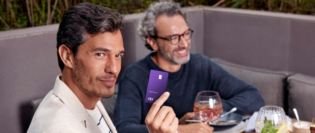 Foto mostra dois homens brancos sentados diante de uma mesa de restaurante. Um deles está no plano de fundo e veste uma blusa azul marinho, usa óculos e tem cabelos grisalhos curtos e barba também grisalha. Outro homem, no plano da frente da foto, está olhando para o lado e segura um cartão Ultravioleta. Ele está com terno cinza e camiseta branca com listras pretas.