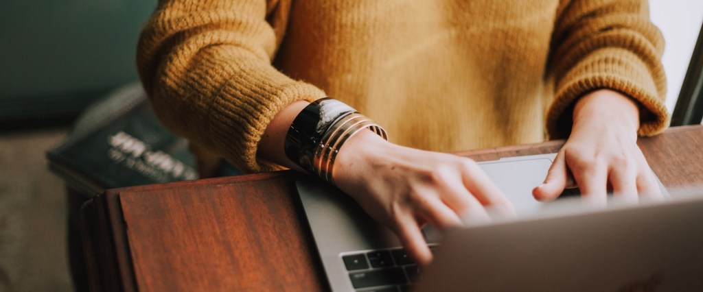 Imagem de uma mulher branca em frente a um notebook digitando alguma coisa. Não vemos o rosto dela. Ela usa uma blusa amarela e pulseiras na mão direita.