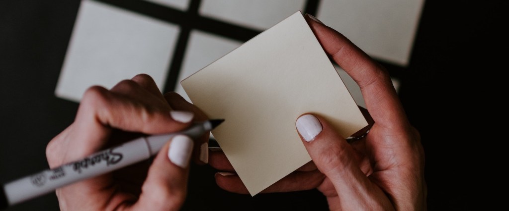 Imagem onde a mão de uma mulher branca segura um post it e uma caneta. Ao fundo, vemos uma mesa preta com mais post its espalhados. As unhas da mulher estão pintadas de branco.