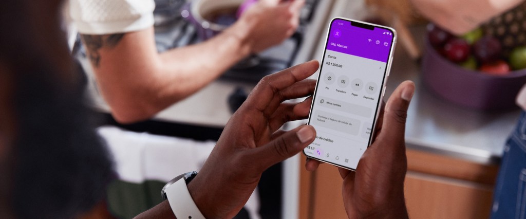 Imagem de um homem negro segurando o celular com a tela inicial do app do Nubank aberta. Ele está em uma cozinha e é possível ver os braços de outras duas pessoas brancas próximas a ele, uma cozinhando e outra organizando um pote com frutas.