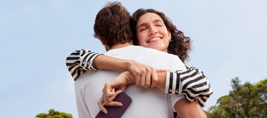 Imagem de um homem e uma mulher se abraçando. Ela está com um celular em mãos e sorrindo. Ele usa camiseta branca e ela uma blusa de manga comprida listrada.