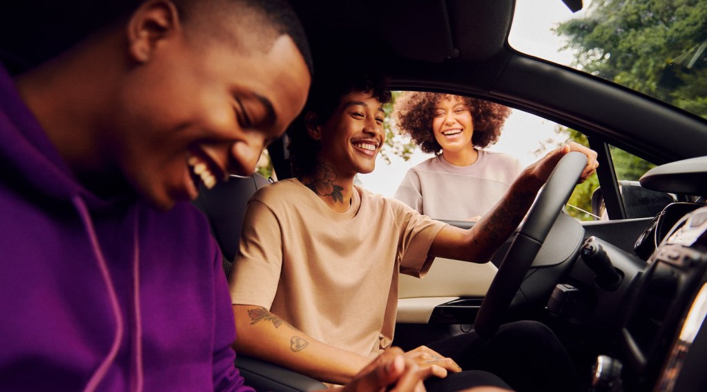 Na foto, um homem dirigindo o carro, um sentado no banco do passageiro e uma mulher do lado de fora, na janela. Todos sorrindo.