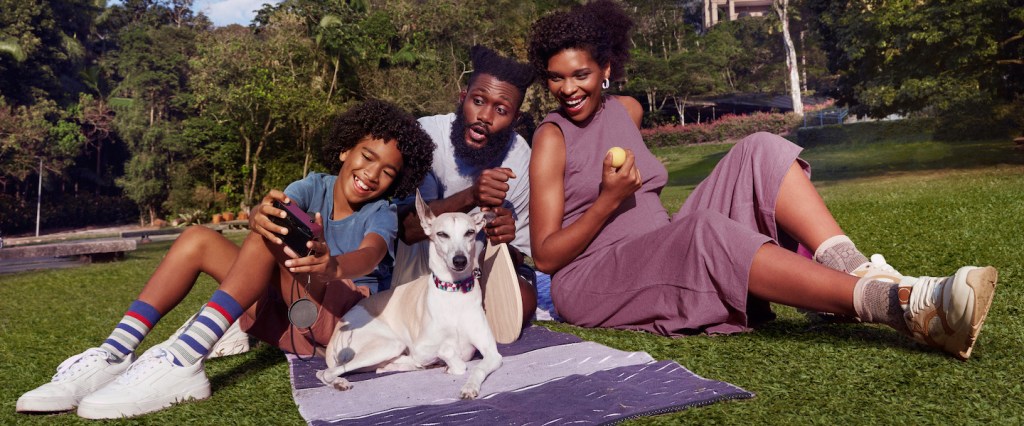 Como funciona o Modo Rua do Nubank? Imagem de uma família negra em um parque. A família é composta por um homem, uma mulher, um menino e um cachorrinho. Eles estão sentados sobre uma canga e assistem algo por um celular, que está nas mãos do menino.