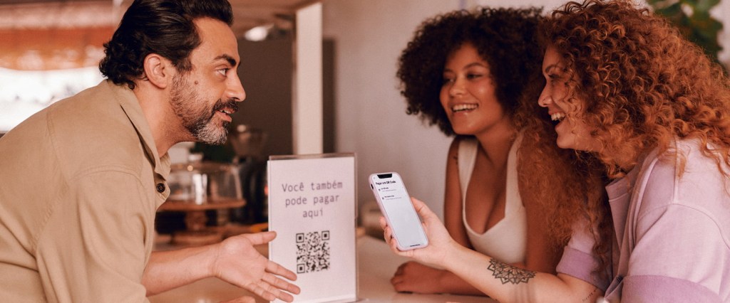 Como gerar QR Code Pix no app do Nubank? Duas mulheres de cabelos cacheados, uma negra e uma ruiva, sorrindo e olhando para a tela do celular enquanto fazem um pagamento via QR Code. Do outro lado do balcão, um homem branco, de cabelos e barba preta, indicando com a mão o código QR Code impresso.