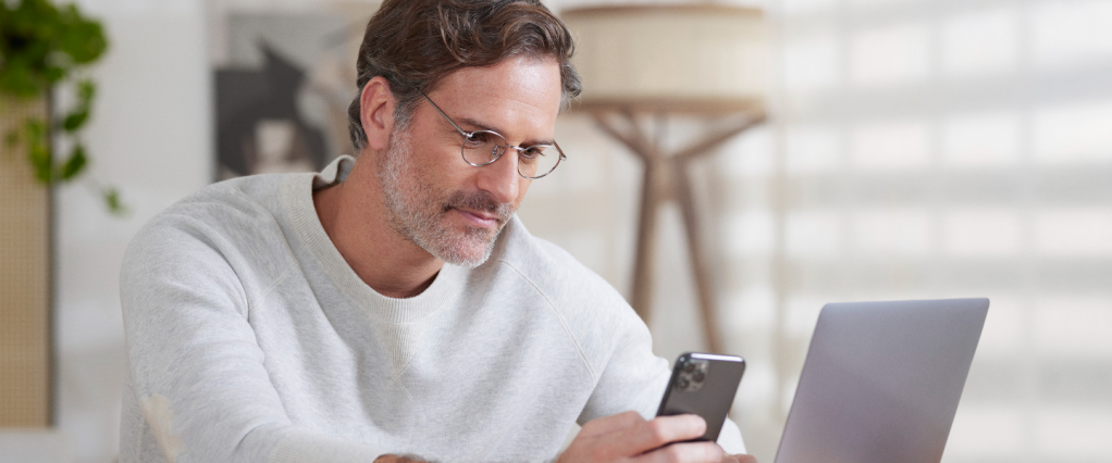 Criptomoedas Nubank: na imagem, uma homem branco, com barba grisalha e camisa branca, está sentado a uma mesa, de frente para um notebook, enquanto olha para o celular, que está segurando com uma das mãos.