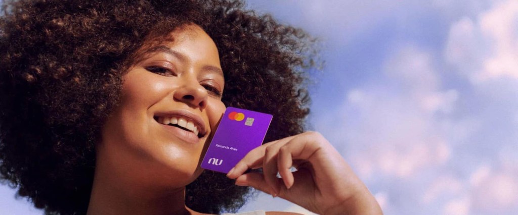 Mulher negra, com cabelos cacheados, sorrindo, ao ar livre, segurando o cartão Nubank próximo ao rosto.