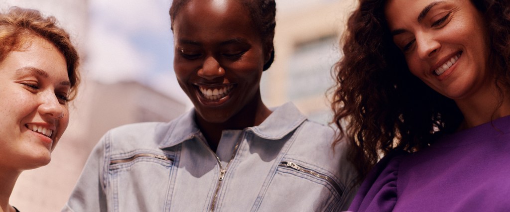 Imagem de três mulheres sorrindo, uma ao lado da outra. Da esquerda para a direita, vemos: uma mulher branca, ruiva, de cabelos cacheados; uma mulher negra, vestindo camisa jeans e com os cabelos trançados pela raiz; uma mulher branca, de cabelo escuro e ondulado, usando uma blusa roxa.