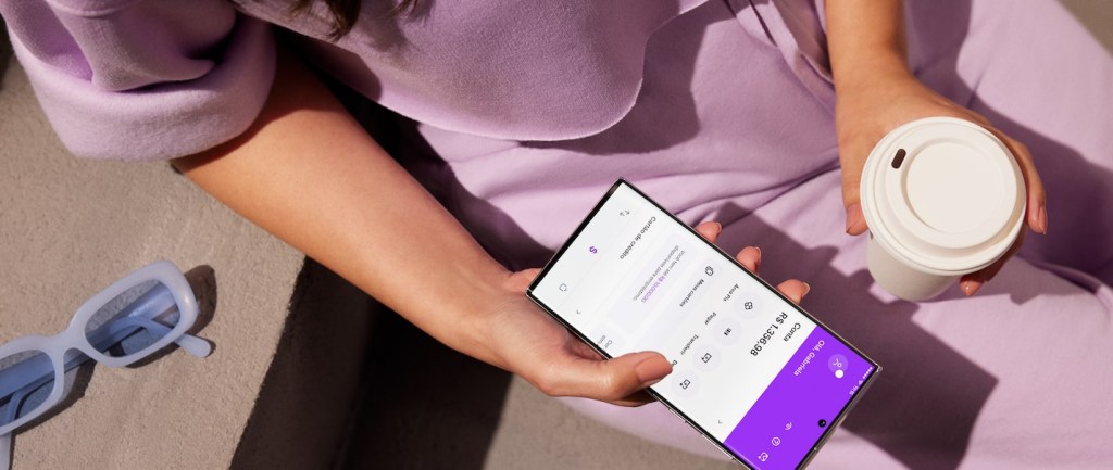 Foto mostra uma mulher vestida toda de roxo claro segurando um celular que mostra o app do Nubank. Na outra mão, ela segura um copo de café e há um óculos ao lado dela.