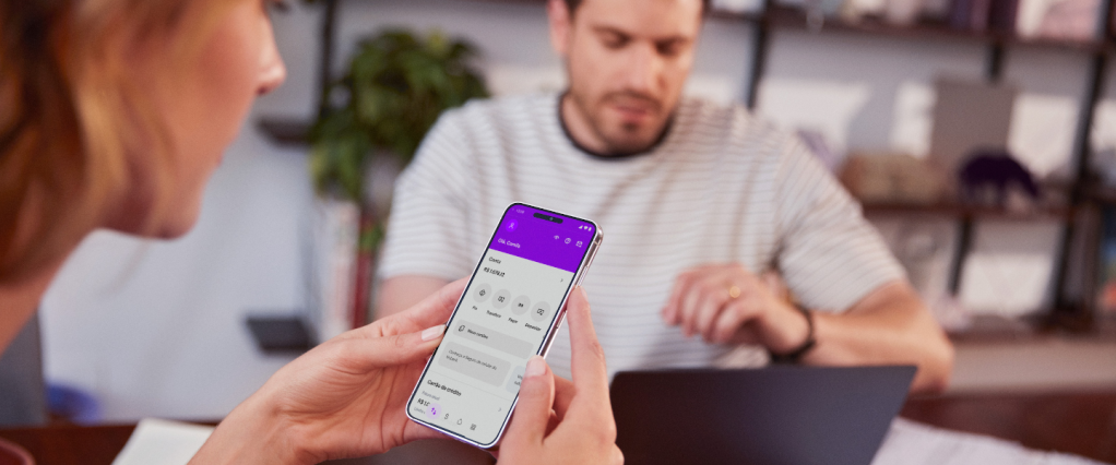 Nubank Acordo Certo: imagem evidencia uma mulher branca, de cabelos loiros, segurando um celular em mãos, com a conta do Nubank na tela. Ao fundo, um branco, tem cabelos pretos curtos e barba. Ele veste uma camiseta listrada branca. Em cima da mesa estão uma caneca, um notebook e vários papéis.