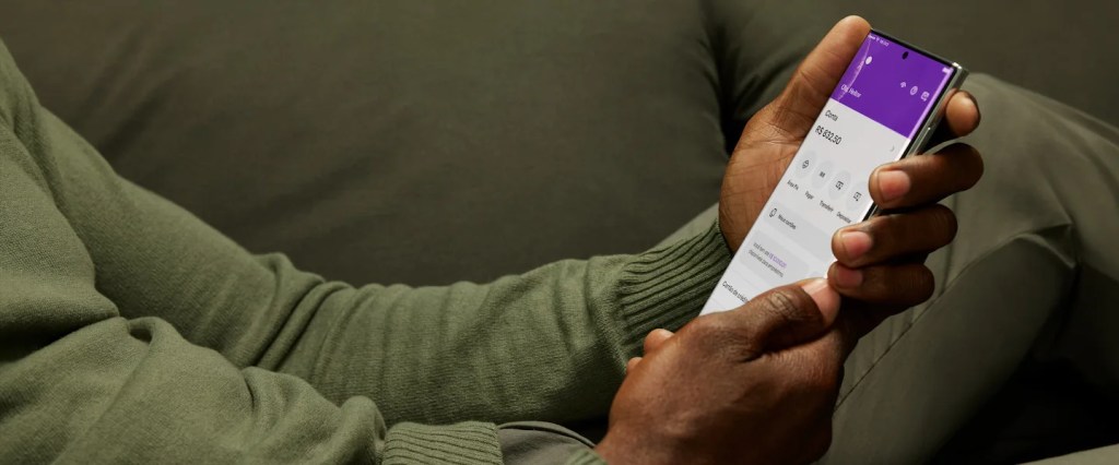 Onde guardar minha reserva de emergência no Nubank? Imagem mostra as mãos de um homem negro vestindo uma camiseta de manga comprida cor verde musgo e segurando um celular na tela inicial do app do Nubank enquanto está sentado em um sofá também na cor verde musgo.