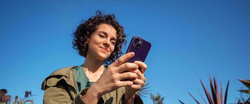 uma mulher sorrindo segurando o celular. De fundo, o céu azul.