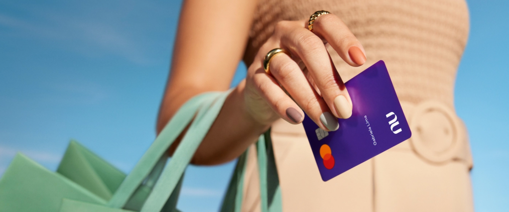 Imagem de uma mulher segurando um cartão de crédito roxinho do Nubank. O nome inscrito no cartão é Gabriela Lima. A mulher tem as unhas pintadas em tons pastéis e segura uma sacola verde pelo antebraço.