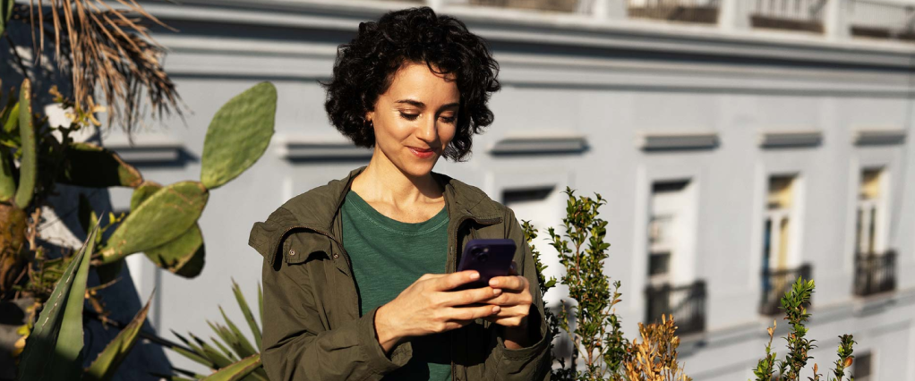 Mulher branca, de cabelos castanhos, curtos e cacheados, olha em direção a um celular roxo que está em suas mãos. Ela está ao ar livre, com um prédio cinza claro ao fundo e plantas ao seu redor. Ela veste uma blusa verde escuro e um casaco em um tom de verde militar.