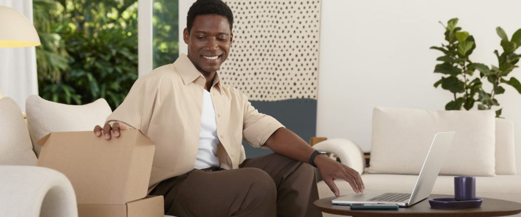 Um homem negro de pele escura, de cabelos bem curtos, vestindo roupas em tons de bege, branco e marrom, está sentado em um sofá ao centro de uma sala. Do lado esquerdo, ele olha para um caixa de papelão, como se fosse uma compra, e, do outro lado, está com o braço esticado em direção a um notebook prata, em cima de uma mesa de centro em madeira. A sala é clara, com plantas e uma bandeira de tecido pendurada ao fundo.