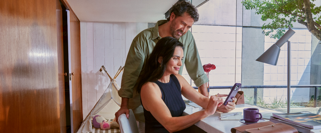 Saldo compartilhado UV: na imagem, um homem e uma mulher brancos estão em um escritório, olhando para um celular com capinha roxa na mão da mulher. Ela está senda em uma mesa, de frente para uma janela e ele está em pé ao seu lado.