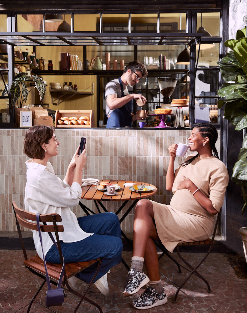 Imagem de duas mulheres conversando, uma de pele clara e cabelos escuros, e a outra negra com tranças. Elas estão tomando café em uma cafeteria. Atrás do balcão, um homem está fazendo café.